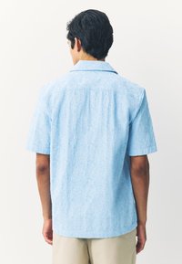 Chemise bleu clair à manches courtes avec motif floral texturé, tissu doux, col classique et ourlet droit, portée avec un short beige.