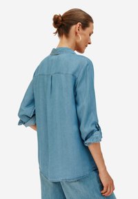 Camicia in denim azzurro chiaro con maniche arrotolate, dettagli con bottoni e piega sul retro. Vestibilità comoda e orlo curvo.