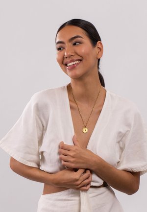 Goldkette mit kreisförmigem Anhänger, getragen über einem hellen, locker sitzenden Leinenoberteil. Glatte Textur, minimalistisches Design.