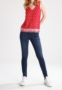 Haut rouge sans manches avec un col en V, doté d'un motif géométrique blanc et d'un ourlet bordé. Porté avec un jean skinny foncé et des baskets blanches.