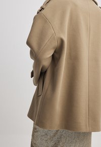 Beżowy płaszcz z wełnianej mieszanki z zaokrąglonymi ramionami, ozdobiony wiązaniem w talii i luźnym krojem, przedstawiony na neutralnym tle.