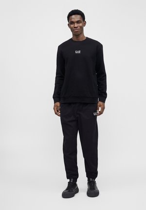 Homme portant un sweat-shirt noir EA7 et un pantalon noir assorti avec des baskets noires, debout devant un fond gris uni.