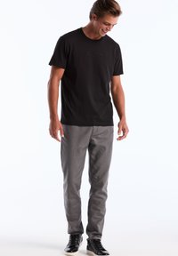 T-shirt noire à manches courtes avec un logo "Original Penguin" texturé, associée à un pantalon gris et des baskets noires, se tenant devant un fond uni.