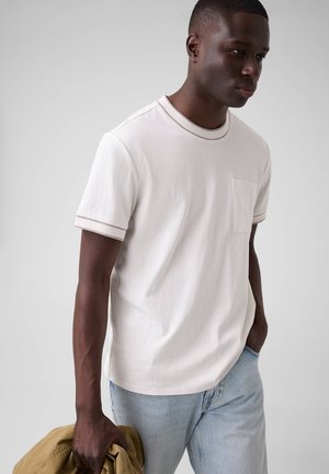 Homme portant un t-shirt blanc à col rond avec poche et un jean bleu clair, tenant une veste beige pliée à ses côtés, sur un fond gris uni.