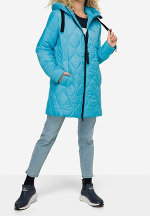 Personne portant un manteau matelassé bleu vif avec capuche, un jean bleu clair et des baskets slip-on bleu foncé, debout avec une jambe en avant.