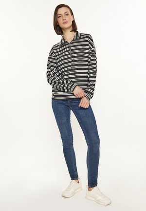 Maglione a righe black and white con colletto e zip fino a metà. Abbinato a jeans blu e sneakers bianche. Sfondo semplice.