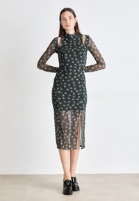 Black mesh dress with green eye pattern, high neck, and cut-out shoulder details. Features a mid-length design with a side slit.