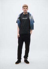 Μαύρο βαμβακερό μπλουζάκι με λευκό κείμενο "CALVIN KLEIN NEW YORK," συνδυασμένο με μαύρη φόρμα και μπλε σκουρόχρωμο κασκόλ. Μαύρα αθλητικά παπούτσια.