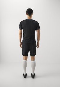 Svart atletisk t-shirt med korta ärmar, i kombination med svarta shorts. Modellen har på sig vita knästrumpor och svarta sportskor.