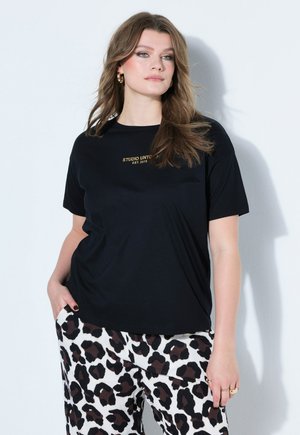 Donna con capelli castani sciolti, che indossa una maglietta nera con la scritta gialla "STUDIO UNTO" e pantaloni bianchi con stampa leopardata, in piedi contro un muro bianco.