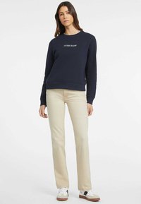 Felpa blu navy con logo "GUESS JEANS", collo rotondo, polsini a costine, abbinata a pantaloni beige chiaro e sneaker bianche con dettagli.