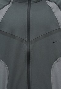 Σκούρο γκρι αθλητικό σακάκι με μαύρο φερμουάρ, ανοιχτό γκρι πλαϊνά πάνελ και μικρό μαύρο λογότυπο Nike στο δεξί στήθος.