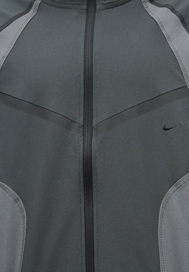 Dunkelgraue Sportjacke mit schwarzem Reißverschluss, hellgrauen Seitenpaneelen und kleinem schwarzem Nike-Swoosh auf der rechten Brust.