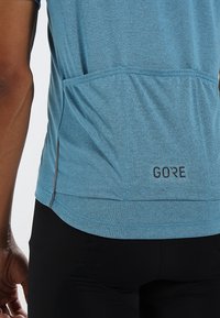 Technické atletické tričko z melírované modré látky. Má zadní kapsu, decentní logo a kontrastní tmavé švy po stranách.