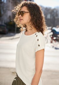 Kobieta z kręconymi włosami i okularami przeciwsłonecznymi w oprawkach w kolorze żółwia, ubraną w białą bluzkę z guziczkami na ramionach i paskowane spodnie na zewnątrz, w słoneczny dzień.