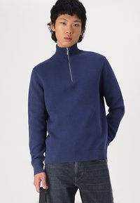 Jūrinis zip-up megztinis, pagamintas iš reljefinio mezgimo audinio. Turi ilgas rankoves, aukštą apykaklę ir prigludusį juosmenį. Suderintas su tamsiomis džinsais.