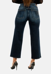 Donkerblauwe, high-waisted jeans met een recht gesneden ontwerp, voorzien van twee achterzakken en contrasterende stiksels. Gemaakt in combinatie met zwarte hoge hakken.