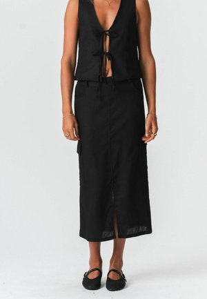 Kvinde iført en sort, ærmeløs top med bindebånd foran, et sort midi-nederdel med høj talje og sorte lukkede sko med ankelstropper.