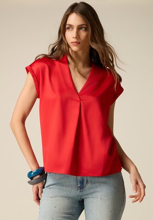 Vrouw met lang bruin haar, gekleed in een mouwloze rode blouse met een V-hals en lichtblauwe spijkerbroek, geaccentueerd met chunky armbanden aan haar linker pols.