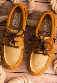 Chaussures bateau en daim beige et marron avec des lacets foncés sur une surface en bois entourée de cordes épaisses beige enroulées.