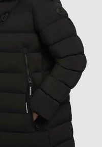Giacca imbottita nera con primo piano di un design trapuntato, tasca con zip e dettaglio del logo circolare sulla manica.