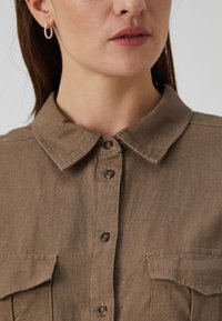 Καφέ πουκάμισο με κουμπιά και διπλωμένο γιακά, δύο τσέπες στο στήθος και ελαφριά υφή. Διαθέτει σκούρα κουμπιά και απλό, κλασικό σχέδιο.