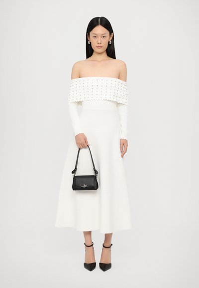 Witte off-shoulder jurk met een textuur bodice en gegolfde randen, gecombineerd met een zwarte leren handtas en puntige zwarte hakken.