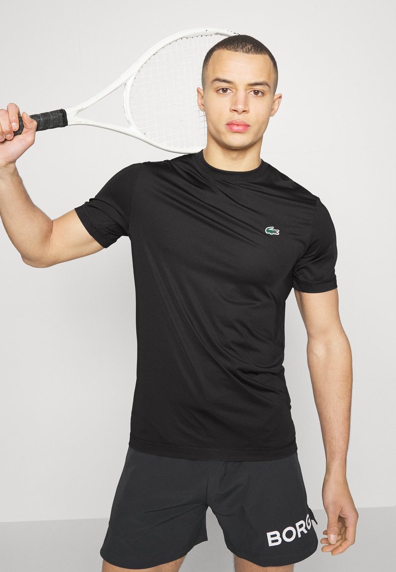 Schwarzes sportliches T-Shirt aus feuchtigkeitsableitendem Material, mit rundem Halsausschnitt und kurzen Ärmeln, mit einem kleinen grünen Logo auf der Brust.