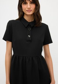 Vestido negro con cuello japonés y mangas cortas, con tres botones con detalles plateados y textura de falda fruncida. Acabado de tela suave.