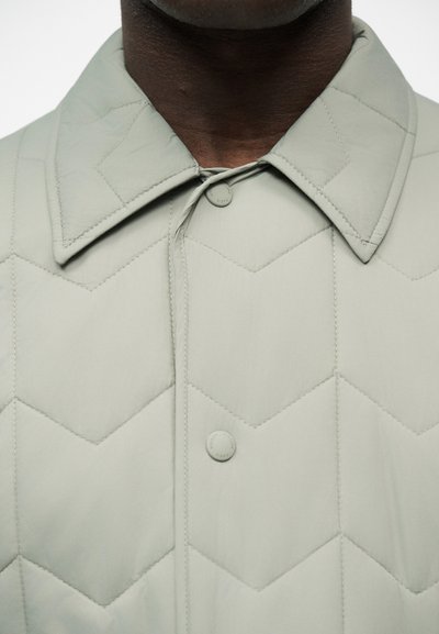 Gros plan d'une veste matelassée gris clair avec des coutures en zigzag, un col pointu et des boutons-pression sur une personne à la peau foncée.