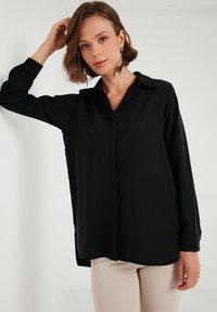 Black long-sleeve button-up shirt with a collar, smooth fabric, and side slits, paired with light beige fitted pants.