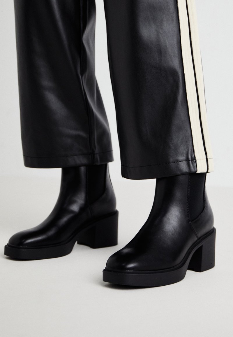 Bottes courtes en cuir noir à talons carrés portées avec un pantalon noir orné de bandes blanches sur les côtés.