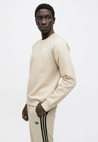 Béžová mikina s okrúhlym výstrihom a ukončenými rukávmi, s malým bielym logom Adidas na hrudi, v kombinácii s rovnakými teplákmi.