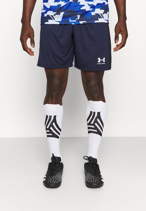 Athlet trägt marineblaue Under Armour-Shorts, weiße knielange Socken mit schwarzem geometrischem Muster und schwarze Fußballschuhe, steht vor einem weißen Hintergrund.