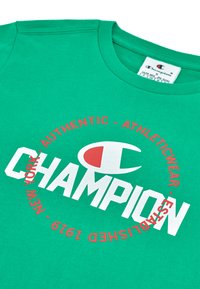 Camiseta de algodón verde con un audaz texto en blanco que dice "CHAMPION" y acentos circulares rojos, con una etiqueta que indica la marca y las instrucciones de cuidado.