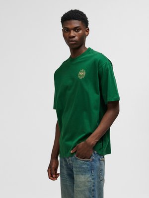 Jonge man draagt een groen T-shirt met een klein rond logo en lichtblauwe spijkerbroek, staand voor een effen lichtgrijze achtergrond.