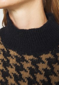 Pletený svetr s vysokým černým límcem, zdobený hnědo-černým vzorem pepitky. Měkká textura s viditelnými vlákny v designu.