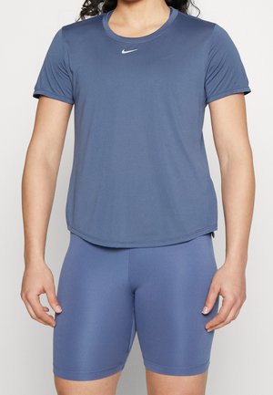 Vrouw draagt een blauw atletisch shirt met korte mouwen en een witte Nike-logo, gecombineerd met nauwsluitende fietsshorts, tegen een effen achtergrond.