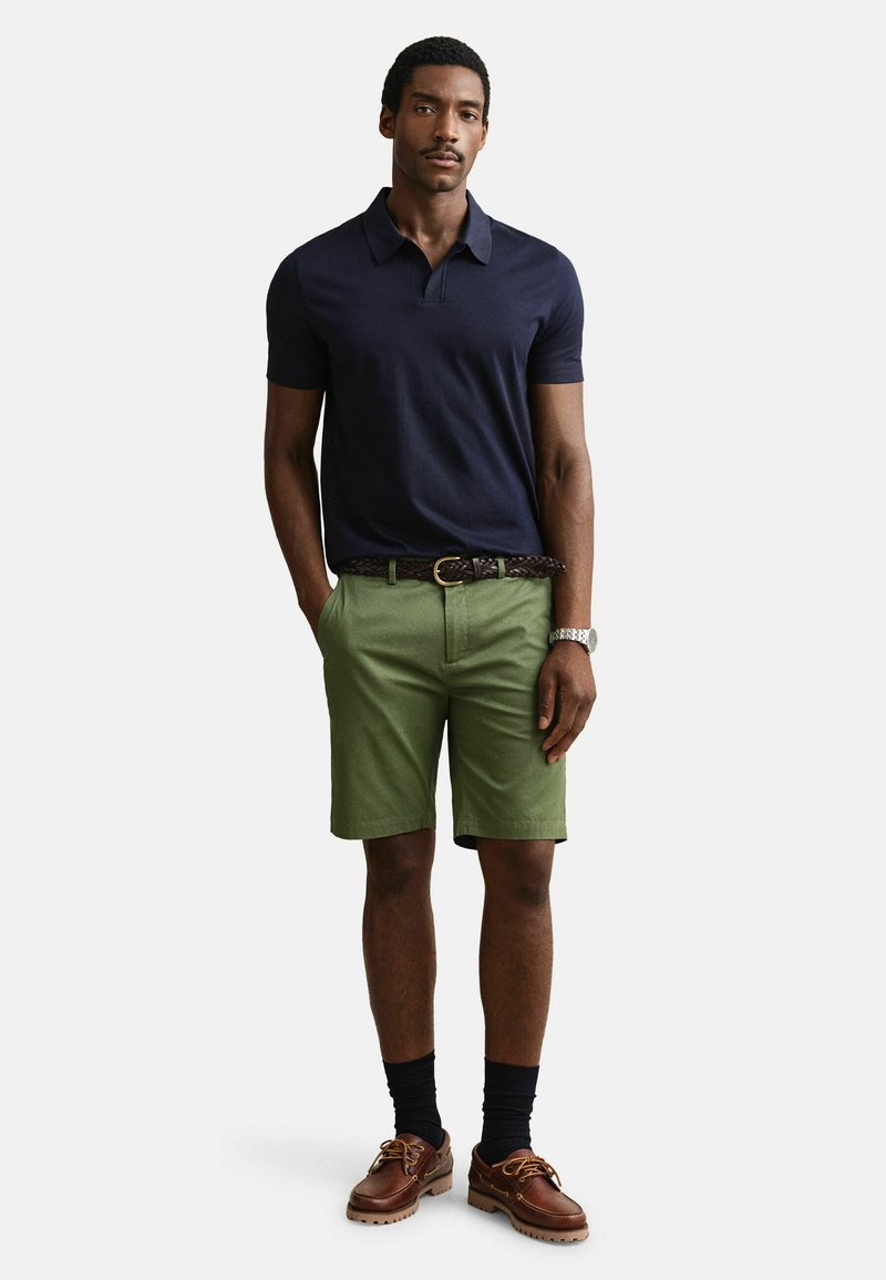Hombre con camiseta polo azul marino, pantalones cortos verde oliva con cinturón trenzado, calcetines negros y zapatos de cuero marrón, de pie con las manos en los bolsillos.
