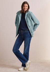 Hellblaue wasserdichte Jacke mit Knopfdetails, getragen über einem marineblauen Sweatshirt, kombiniert mit blauen Jeans und weißen Sneakern. Glatte Texturen sichtbar.