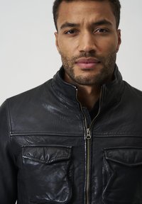 Svart läderjacka med hög krage, dragkedja fram och två bröstfickor. Ytan har en lätt texturerad finish.