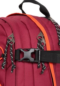 Punainen kangaskassi, jossa on oranssit yksityiskohdat, mustat hihnat ja metalliset vetoketjun vedin. Mukana muovinen solki ja koristeellinen nyöri.