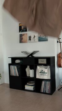 Μαύρη ξύλινη βιβλιοθήκη με ανοιχτές θήκες που εκθέτουν βιβλία, μια βάζο, ρούχα και ένα ηχείο. Λείο επιφάνεια και μινιμαλιστικός σχεδιασμός.