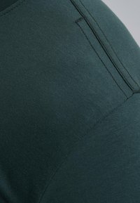 Tamnozeleni prilagođeni odjevni predmet s glatkom teksturom, koji ima bočnu džep i suptilne detalje šivanja. Tkanina izgleda rastezljivo.