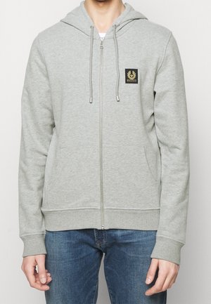 Mand iført lysegrå zip-up hættetrøje med snore og lille sort patch-logo, kombineret med blå jeans.