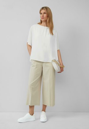 Haut blanc à manches courtes, pantalon large beige et baskets blanches. Le modèle tient un sac rond blanc avec une bandoulière en chaîne dorée.