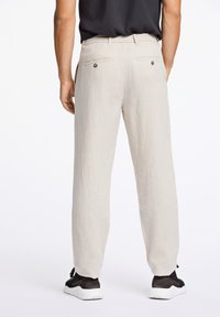 Lichte beige broek van een gestructureerde stof, met een rechte snit, twee achterzakken met knopen en een elastische tailleband, gecombineerd met zwarte schoenen.