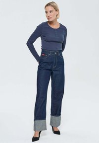 Femme debout avec les mains derrière le dos, portant une chemise longue à manches navy, un jean noir retroussé et des chaussures à talons hauts noires sur fond blanc.