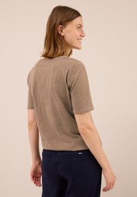 Kvinde smilende, vendt mod højre, iført en brun kortærmet skjorte og mørkeblå bukser foran en ensfarvet beige baggrund.