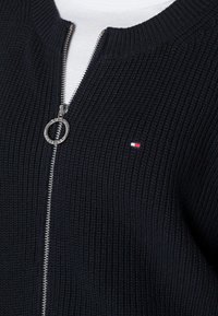 Černá pletená bomber bunda se stříbrným zipem a kulatým táhlem. Malá nášivka s červeno-bílou-modrou logem na levé hrudi.